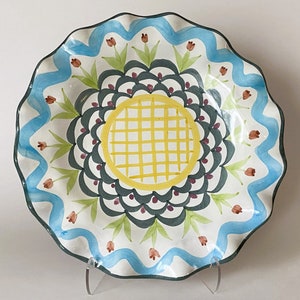 May include: A white ceramic serving dish with a scalloped edge. The dish features a blue and green floral design with a yellow center.