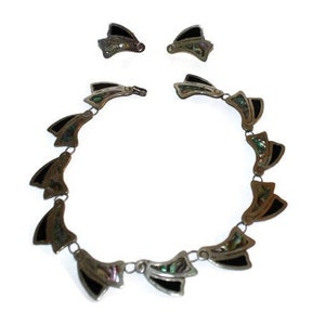 May include: A silver necklace and earrings set with black and green abalone shell accents. The necklace is made of a chain with a series of pendant-like pieces that are shaped like a fan.