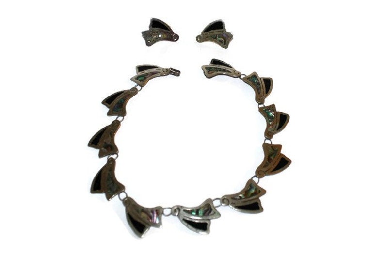 May include: A silver necklace and earrings set with black and green abalone shell accents. The necklace is made of a chain with a series of pendant-like pieces that are shaped like a fan.