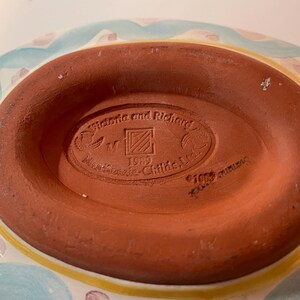 May include: The underside of a ceramic dish featuring a terracotta-colored base with an oval stamp. The stamp reads Victoria and Richard, MacKenzie-Childs, Ltd., 1989, and includes a square logo with diagonal lines. The rim shows blue and yellow paint.