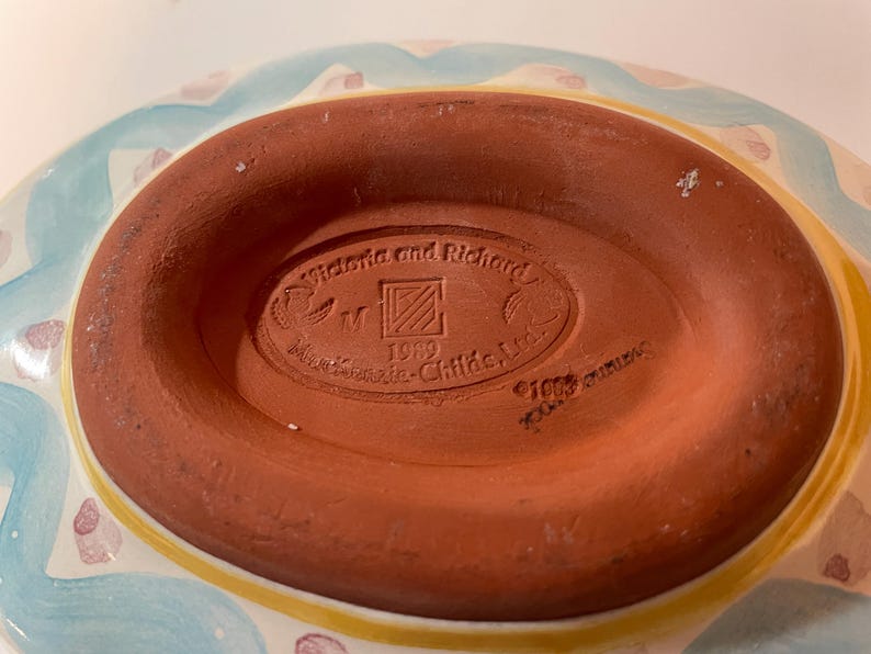 May include: The underside of a ceramic dish featuring a terracotta-colored base with an oval stamp. The stamp reads Victoria and Richard, MacKenzie-Childs, Ltd., 1989, and includes a square logo with diagonal lines. The rim shows blue and yellow paint.
