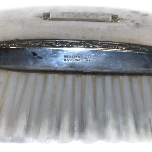 May include: A silver-plated hair brush with a white bristle brush. The handle has an engraved design and the words "Webster Sterling" on the bottom.