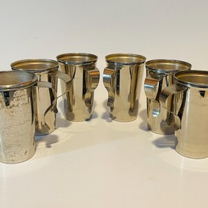 May include: Six silver-colored metal mugs with handles. The mugs have a cylindrical shape with a slightly flared top. The handles are a curved, open design. The mugs are arranged in a group on a white surface.