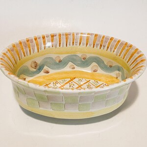 May include: An oval ceramic bowl featuring a yellow rim with vertical brown stripes. The interior displays a light blue wavy line, brown dots, and a central yellow section with a cross-hatched pattern. The exterior has a light green checkerboard.