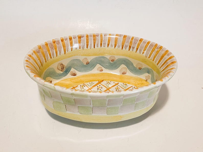 May include: An oval ceramic bowl featuring a yellow rim with vertical brown stripes. The interior displays a light blue wavy line, brown dots, and a central yellow section with a cross-hatched pattern. The exterior has a light green checkerboard.
