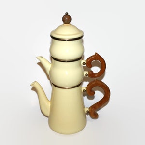 May include: A white enamel coffee pot with a brown wooden handle and a gold trim. The pot has a tiered design with a small lid on top.