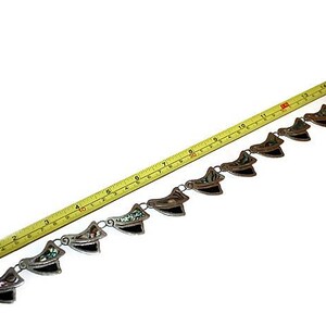 May include: A silver chain necklace with a series of decorative pendants. Each pendant is made of silver and features a black and green abalone shell inlay. The necklace is measured with a tape measure, showing it is approximately 15 inches long.