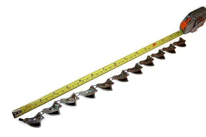 May include: A silver chain necklace with a series of decorative pendants. Each pendant is made of silver and features a black and green abalone shell inlay. The necklace is measured with a tape measure, showing it is approximately 15 inches long.