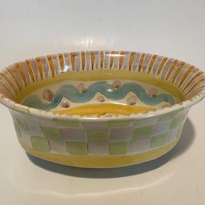 May include: An oval ceramic bowl featuring a hand-painted exterior with a light green and white checkered pattern above a solid yellow base. The interior rim displays vertical orange and grey stripes, while the center shows a wavy blue line.