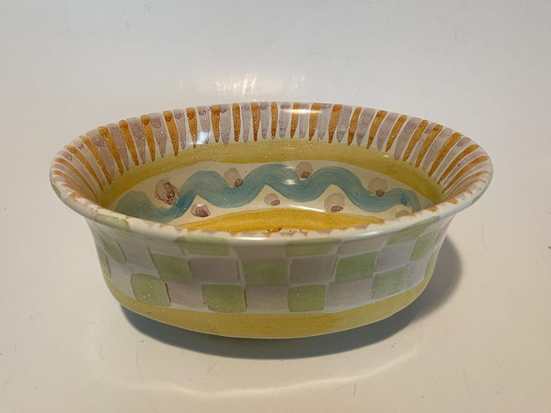 May include: An oval ceramic bowl featuring a hand-painted exterior with a light green and white checkered pattern above a solid yellow base. The interior rim displays vertical orange and grey stripes, while the center shows a wavy blue line.