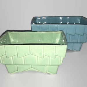 May include: Two ceramic planters, one light green and one light blue, with a geometric design. The planters are rectangular with a slightly curved bottom and have a textured surface.