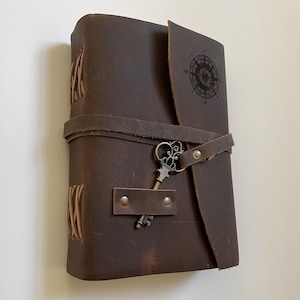 May include: A brown leather journal with a compass design and a key charm. The journal has a strap closure and is bound with twine.
