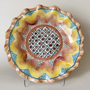 May include: A ceramic serving bowl with a scalloped edge and a colorful hand-painted design. The bowl features a central motif of a geometric pattern in shades of brown, green, and white, surrounded by a ring of yellow, orange, and blue.