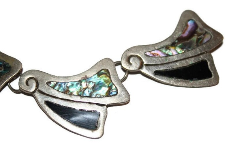 May include: A silver necklace with a geometric design featuring abalone shell and black accents. The necklace is made of multiple pieces that are connected together.
