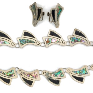 May include: A silver necklace and earrings set with black and iridescent abalone shell accents. The necklace is made of a chain of connected abalone shell and black accents in a geometric shape.