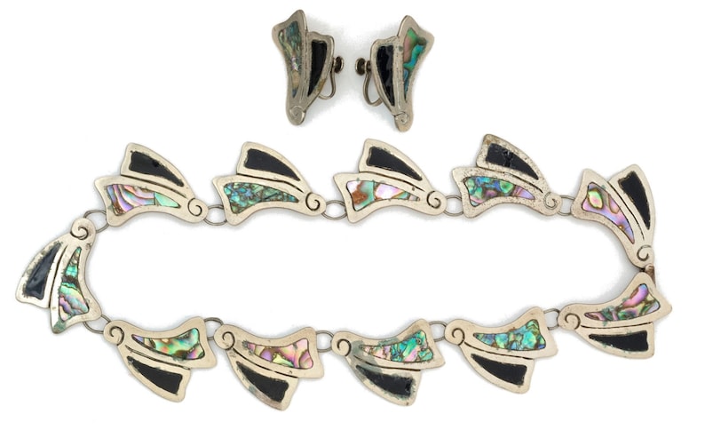 May include: A silver necklace and earrings set with black and iridescent abalone shell accents. The necklace is made of a chain of connected abalone shell and black accents in a geometric shape.