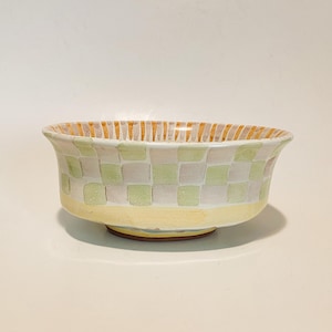 May include: A ceramic bowl featuring a checkered pattern of light green and white squares on the exterior. The base is a solid pale yellow, while the interior rim displays vertical orange and white stripes. The bowl has a flared, rounded shape.