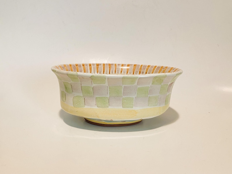 May include: A ceramic bowl featuring a checkered pattern of light green and white squares on the exterior. The base is a solid pale yellow, while the interior rim displays vertical orange and white stripes. The bowl has a flared, rounded shape.