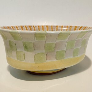 May include: A ceramic bowl featuring a hand-painted checkerboard pattern in soft shades of sage green and pale lavender. The base is a solid band of yellow, while the interior rim is decorated with vertical orange and white stripes.