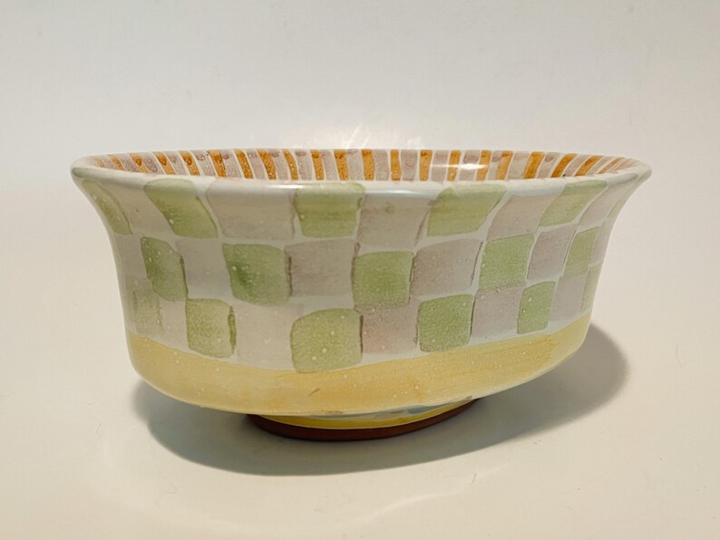 May include: A ceramic bowl featuring a hand-painted checkerboard pattern in soft shades of sage green and pale lavender. The base is a solid band of yellow, while the interior rim is decorated with vertical orange and white stripes.