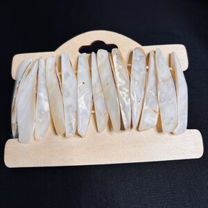 May include: A close-up of a white shell bracelet with a wooden display card. The bracelet is made of many small, flat, white shells that are arranged in a row.