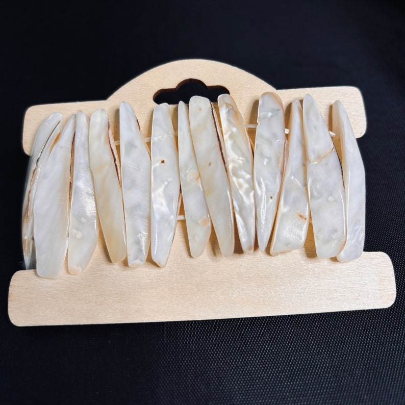 May include: A close-up of a white shell bracelet with a wooden display card. The bracelet is made of many small, flat, white shells that are arranged in a row.