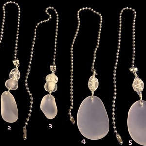May include: Six decorative ceiling fan pull chains are displayed. Each chain has a silver beaded chain, clear glass beads, and a light purple, oval-shaped pendant. The pendants vary in shape and size.