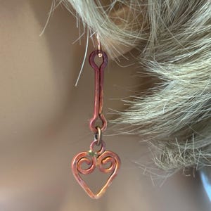 May include: A pair of copper earrings with a heart-shaped dangle. The earrings are a reddish-brown color and have a wire-wrapped design.