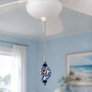 Könnte beinhalten: Eine Deckenventilator-Zugkette mit einer silberfarbenen Metallkugelkette, die an einem dekorativen Anhänger befestigt ist. Der Anhänger besteht aus einem weiß-blauen, spiralförmigen Ammoniten-Fossil-Anhänger, akzentuiert durch zwei dunkelblau schimmernde Perlen.