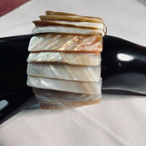 May include: A multi-layered bracelet made of iridescent mother-of-pearl shell pieces. The bracelet is made of a variety of shades of white, cream, and brown.