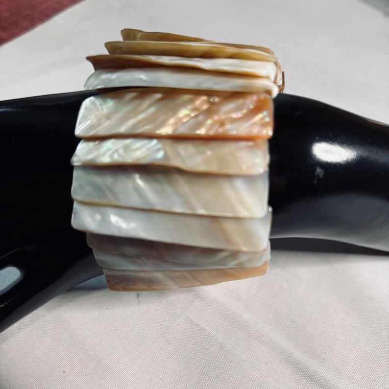 May include: A multi-layered bracelet made of iridescent mother-of-pearl shell pieces. The bracelet is made of a variety of shades of white, cream, and brown.