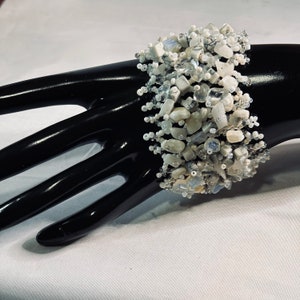 May include: A white and gray beaded bracelet with a cluster of white and gray stones. The bracelet is on a black mannequin hand.