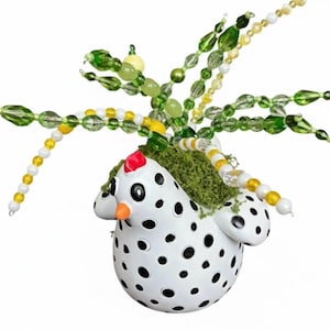 Pode incluir: Uma figura de galinha de cerâmica branca com bolinhas pretas, uma crista vermelha e um bico laranja. A galinha tem as costas verde musgo e é adornada com contas em tons de verde, amarelo e branco.