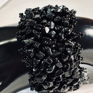 May include: A black beaded bracelet with black gemstone chips. The bracelet is made of small, irregular shaped black stones and black beads.