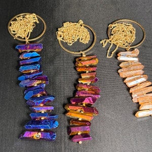 Könnte beinhalten: Drei goldene Halsketten mit Kristallanhängern. Die Anhänger bestehen aus irisierenden, facettierten und gestapelten Kristallteilen. Die Kristalle sind in Blautönen, Lila, Orange und Rosa.