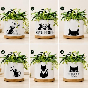 May include: A set of six white cylindrical ceramic planters with bamboo bases, each containing a green beaded plant. The planters feature black cat-themed graphics, including silhouettes, paw prints, and text like Cat Mom and Judging You Silently.