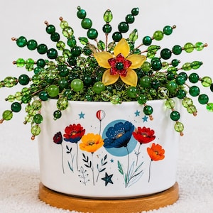 May include: A white ceramic planter with a floral print featuring red, yellow, and blue flowers, stars, and a balloon. It contains a decorative wire sculpture with numerous green beads and a central yellow and red flower-shaped ornament.