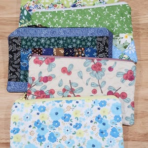 May include: A collection of four rectangular zippered pouches made of quilted fabric. The designs include a blue and white floral print, a cream pouch with red berries, a blue patchwork pattern, and a green fabric with white bows.