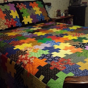 Puede incluir: Un edred&oacute;n colorido y dos fundas de almohada a juego con un patr&oacute;n de rompecabezas. El edred&oacute;n presenta una variedad de patrones y colores de tela, incluyendo rojo, naranja, amarillo, verde, azul y morado. La ropa de cama est&aacute; sobre una estructura de cama de madera.