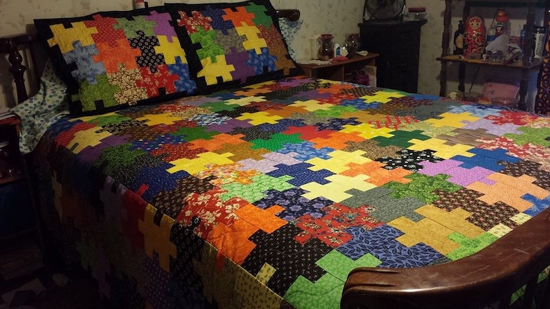 Puede incluir: Un edred&oacute;n colorido y dos fundas de almohada a juego con un patr&oacute;n de rompecabezas. El edred&oacute;n presenta una variedad de patrones y colores de tela, incluyendo rojo, naranja, amarillo, verde, azul y morado. La ropa de cama est&aacute; sobre una estructura de cama de madera.