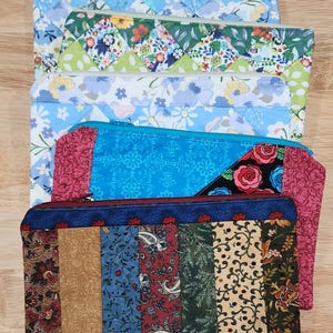 May include: A stack of five rectangular fabric zipper pouches featuring various patterns. The designs include blue and white floral prints, a green diamond patchwork, a bright blue floral print, and a multi-colored vertical striped pouch.