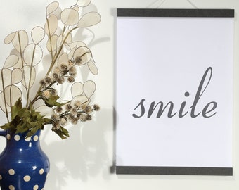 May include: A white framed poster with the word "smile" printed in black cursive script. The poster is hanging on a white wall next to a blue and white polka dot vase with a dried flower arrangement.