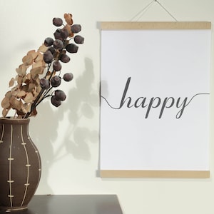 Op de afbeelding: Een witte geframed poster met het woord "happy" geschreven in cursief op een witte achtergrond. De poster hangt aan een witte muur naast een bruine vaas met gedroogde takken en bessen.