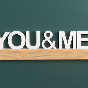 May include: White wooden letters spelling "YOU & ME" are displayed on a light brown wooden shelf against a teal green background.