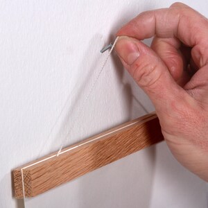 May include: A wooden picture hanging rail with a white string attached to a nail in a white wall. A hand is holding the string to hang the rail.