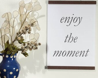 May include: A white poster with a brown wooden frame hangs on a white wall. The poster has the text "enjoy the moment" written in cursive script. A vase with blue and white polka dots holds a dried flower arrangement with white papery leaves.