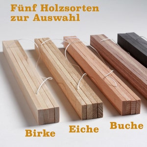 May include: Five wooden strips of different wood types, including birch, oak, beech, and walnut, are stacked and tied together with string. The wood strips are arranged from left to right in order of their color, with the lightest wood on the left and the darkest wood on the right. The wood strips are labeled with their respective wood types in German.