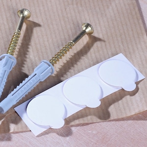 Puede incluir: Dos tacos de pared de pl&aacute;stico blanco con tornillos dorados y tres tacos adhesivos de pared blancos sobre un fondo de papel marr&oacute;n.