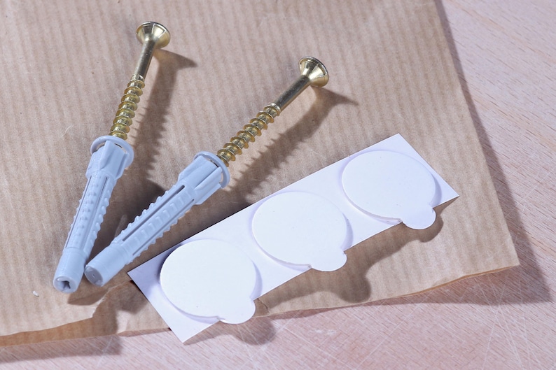Puede incluir: Dos tacos de pared de pl&aacute;stico blanco con tornillos dorados y tres tacos adhesivos de pared blancos sobre un fondo de papel marr&oacute;n.
