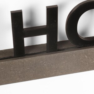 Puede incluir: Letras de madera negras que deletrean "HOME" montadas sobre una base de madera marr&oacute;n.